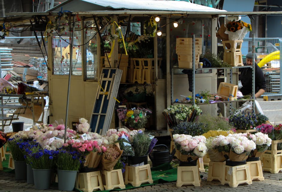 An outdoor flower stall with a variety of fresh floral arrangements displayed on beige plastic crates and black buckets. The flowers include pink peonies, white roses, purple statice, and blue cornflo