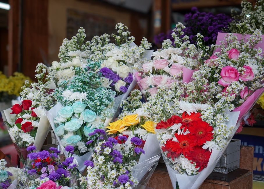 An array of fresh floral bouquets displayed on a wooden surface outside a flower shop. The arrangements feature a variety of flowers including pink roses, red gerberas, yellow roses, white lisianthus,