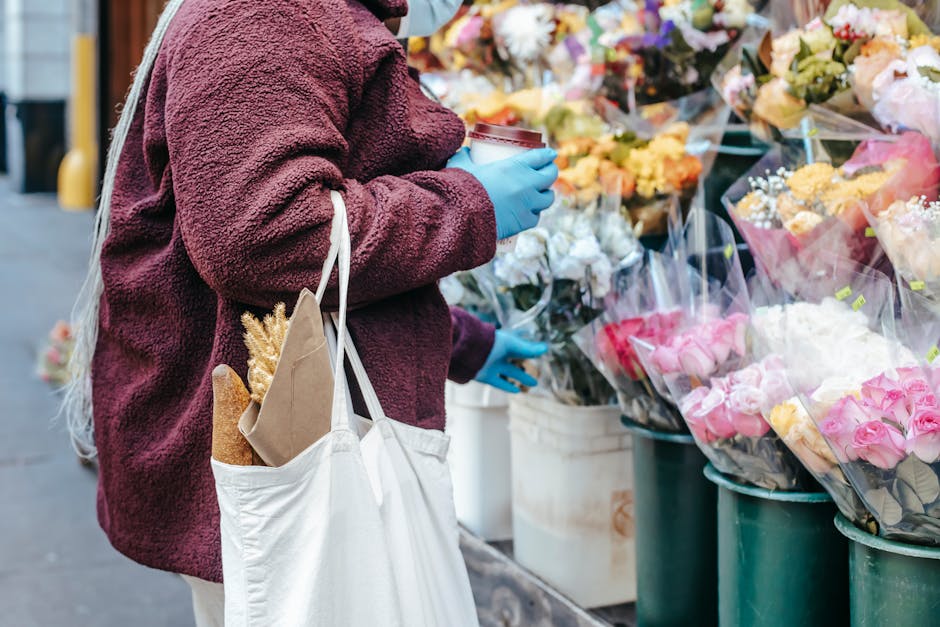 A person wearing a maroon, textured fleece jacket and light-colored pants is standing at an outdoor flower stall, holding a cup and wearing blue gloves. They carry a white reusable tote bag with a bag